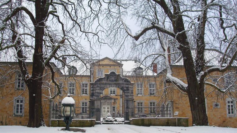 Ein historisches Gebäude im Schnee, umgeben von kahlen Bäumen. Die Atmosphäre ist ruhig und winterlich.