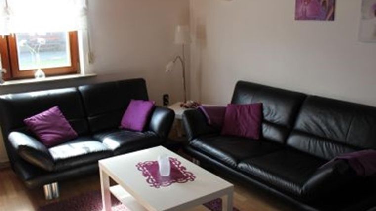 Ein gemütliches Wohnzimmer mit zwei schwarzen Sofas und lila Kissen. Ein kleiner Tisch steht in der Mitte, auf dem eine Kerze steht.