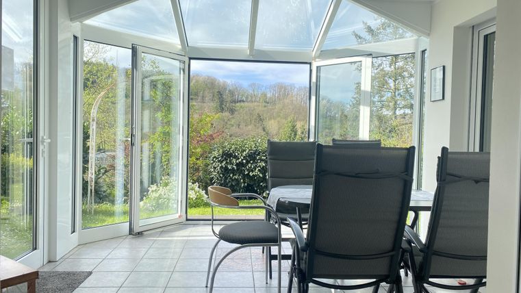 Ein gemütlicher Wintergarten mit offenen Fenstern. Im Hintergrund sind grüne Pflanzen und eine ruhige Landschaft zu sehen.
