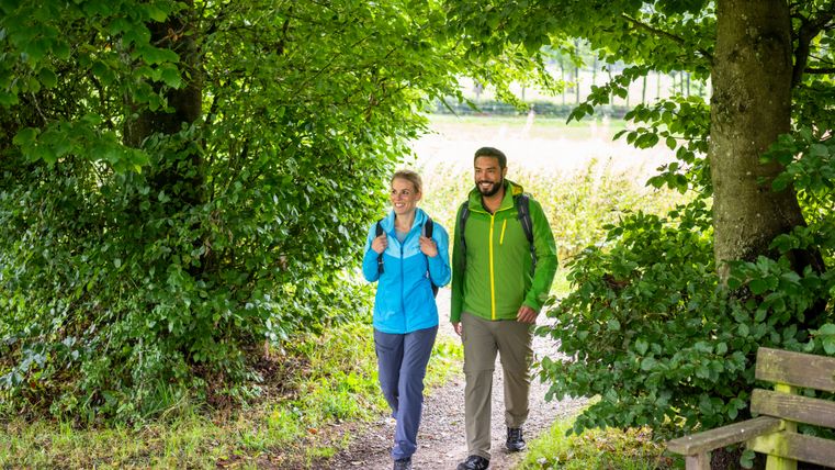 Zwei Wandernde mit Rucksäcken gehen auf einem schmalen Waldweg unter dichtem, grünem Blätterdach entlang. Rechts steht eine hölzerne Bank, im Hintergrund öffnet sich der Blick ins helle Licht.