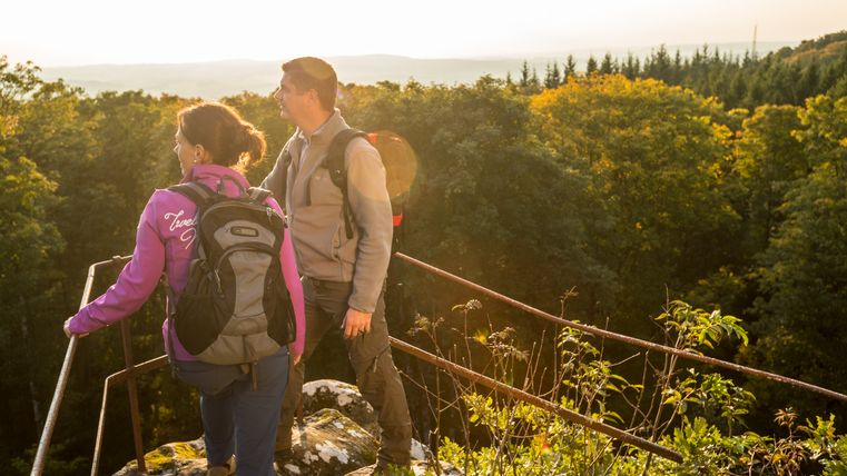Zwei Wanderer stehen auf einem Aussichtspunkt mit Blick auf einen Wald bei Sonnenuntergang.