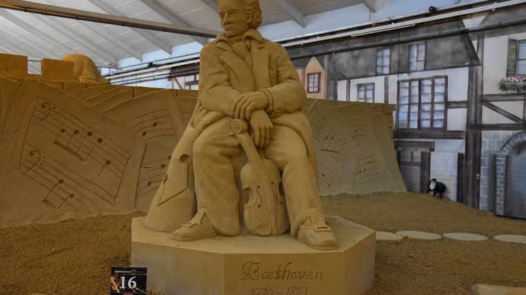 Eine Sandskulptur eines sitzenden Mannes mit einem Instrument. Im Hintergrund sind Sand und eine Nachbildung einer historischen Stadt zu sehen.
