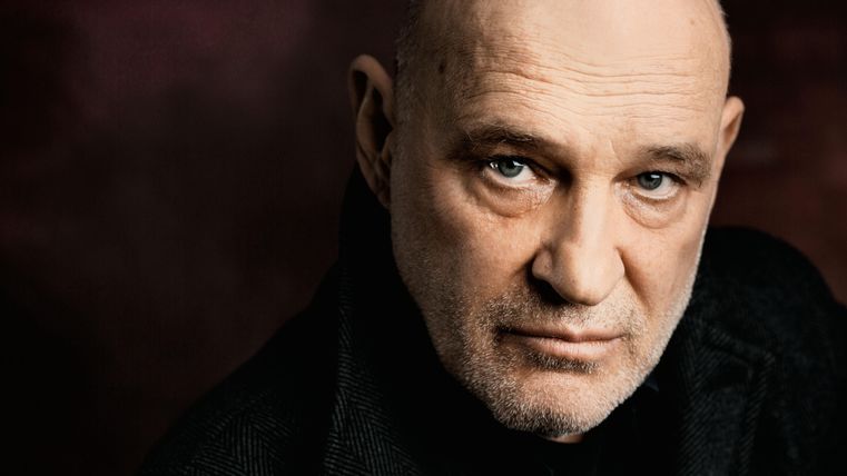 Ein älterer Mann mit Glatze und ernstem Gesichtsausdruck schaut direkt in die Kamera. Der Hintergrund ist dunkel und unscharf, was den Fokus auf sein Gesicht lenkt.