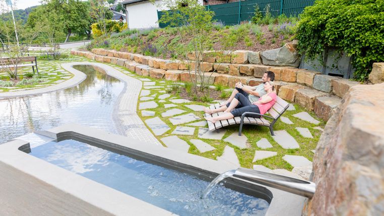 Ein ruhiger Garten mit einem fließenden Wasserlauf und bequemen Liegen. Zwei Personen entspannen sich in der Natur.