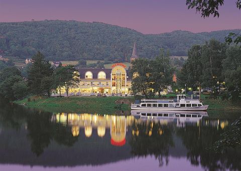 Eine beeindruckende Ansicht entlang eines ruhigen Flusses mit einem beleuchteten Gebäude und einem Schiff im Vordergrund. Die sanften Hügel im Hintergrund schaffen eine idyllische Atmosphäre bei Dämmerung.