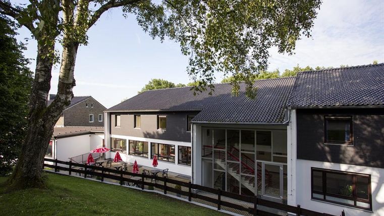Ein modernes Gebäude mit einem großen Fenster und einem einladenden Eingangsbereich. Umgeben von Bäumen und einem grünen Rasen bietet es eine entspannte Atmosphäre.