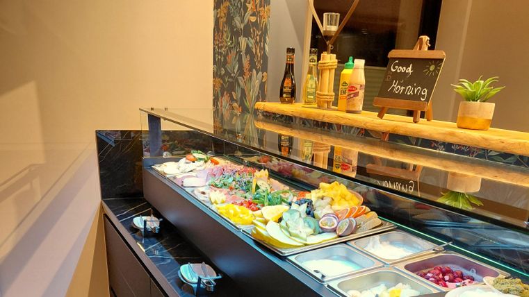 Ein Buffet mit einer Auswahl an frischem Obst, Gemüse und verschiedenen Beilagen. Dazu liegen Flaschen mit Saucen und eine kleine Tafel mit der Aufschrift "Guten Morgen".