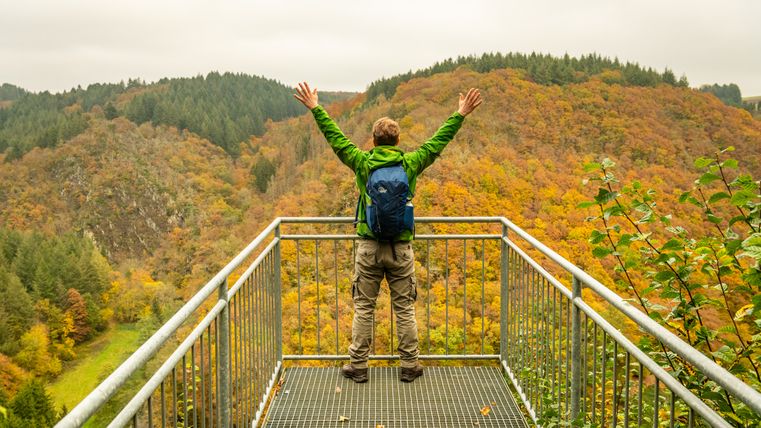 Person auf einer Aussichtsplattform in der Eifel mit ausgestreckten Armen, umgeben von herbstlichen Wäldern.
