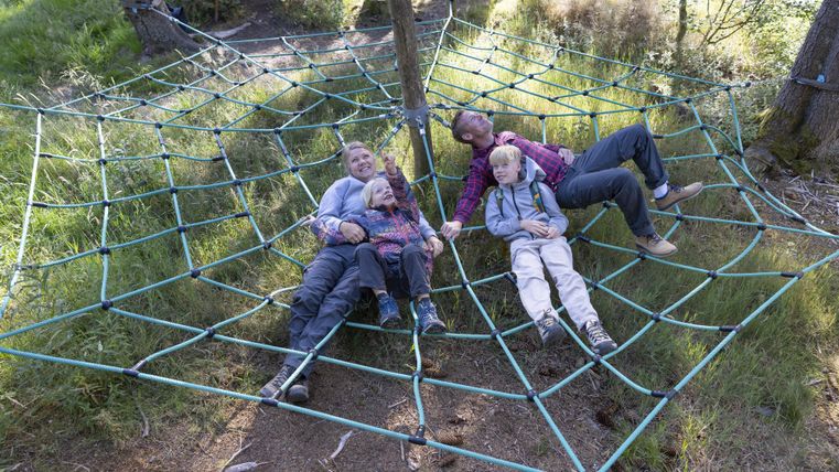 Eine Gruppe von vier Personen liegt entspannt in einem großen Netz aus Seilen in der Natur. Die Umgebung ist grün und freundlich, ideal für Familienaktivitäten.