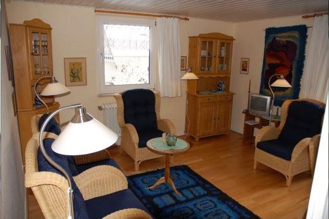 Ein gemütliches Wohnzimmer mit Rattanmöbeln und einem blauen Teppich. Die Wände sind hell, und es gibt einen Fernseher sowie dekorative Elemente.