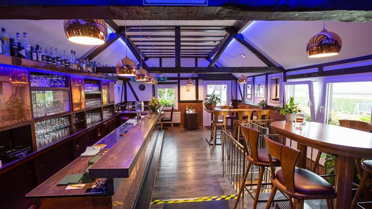 Eine moderne Bar mit Holzmöbeln und stilvollen Beleuchtungselementen. Im Hintergrund sind Fenster mit Blick ins Freie und Getränkesortiment sichtbar.