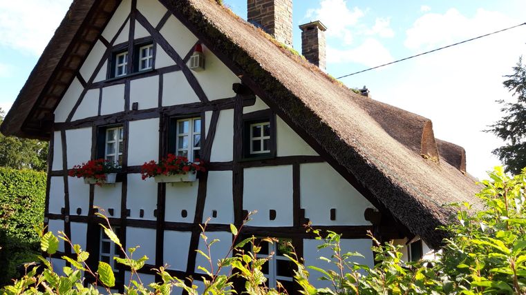 Ein traditionelles Fachwerkhaus mit einem Reetdach und bunten Blumenkästen. Das Gebäude steht in einer grünen Umgebung unter blauem Himmel.