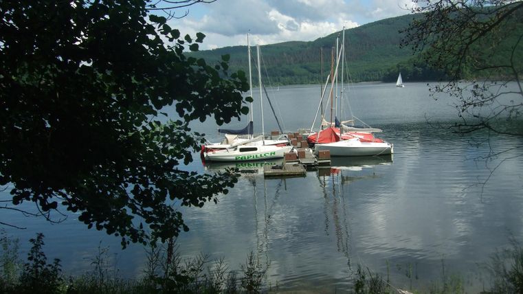 Eine ruhige Seenlandschaft mit mehreren Segelbooten im Wasser. Im Hintergrund sind sanfte Hügel und ein teilweise bewölkter Himmel zu sehen.