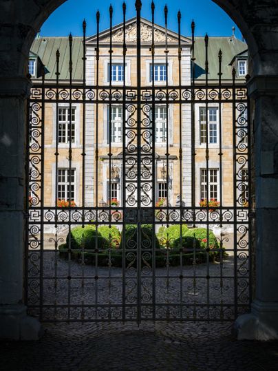 Ein historisches Gebäude, das durch ein schmiedeeisernes Tor sichtbar ist. Der Himmel ist klar und blau, und der Innenhof ist grün bepflanzt.