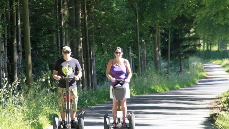 Zwei Personen fahren auf Segways entlang eines Waldwegs. Die Umgebung ist grün und sonnig.