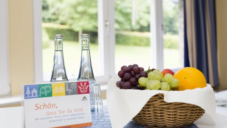 Ein Tisch mit zwei Flaschen Wasser und einer Obstschale, die Trauben, eine Orange und Äpfel enthält. Im Hintergrund sind große Fenster, die auf einen grünen Garten blicken.