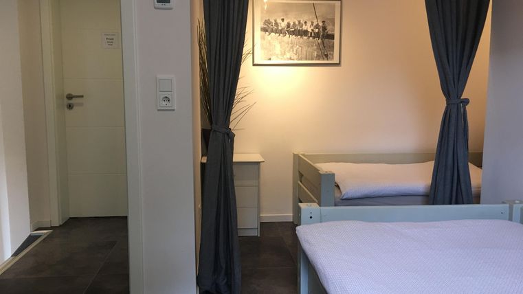 Ein einfaches Zimmer mit zwei Betten und einem Fenster. Vorhänge getrennen den Raum, und an der Wand hängt ein Schwarz-Weiß-Foto.