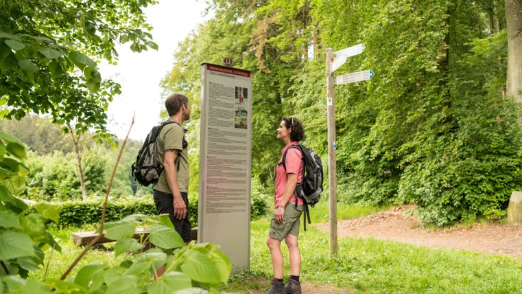 Zwei Wanderer stehen vor einem Informationsschild im Weisshauswald, umgeben von grüner Natur.
