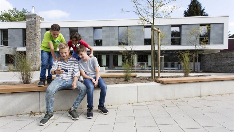 Vier Kinder sitzen und stehen in einer modernen Umgebung. Sie schauen gemeinsam auf ein Handy und wirken dabei interessiert und unterhalten.