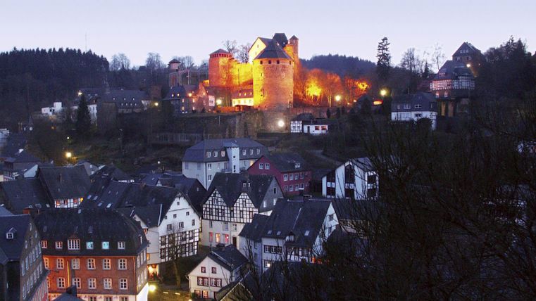 Eine malerische Stadt mit historischen Gebäuden unter einem klaren Himmel. Im Hintergrund leuchtet eine Burg in warmem Licht.