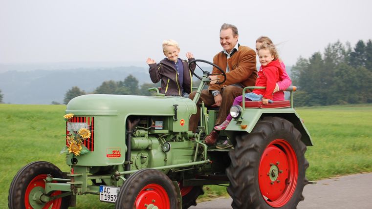 Ein Mann fährt mit zwei Kindern auf einem alten Traktor über einen Feldweg. Die Landschaft im Hintergrund ist grün und ländlich.