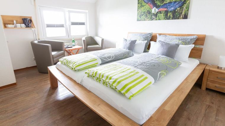 Ein modernes Schlafzimmer mit einem großen Doppelbett und bunten Kissen. In der Ecke stehen zwei gemütliche Sessel und ein Tisch.