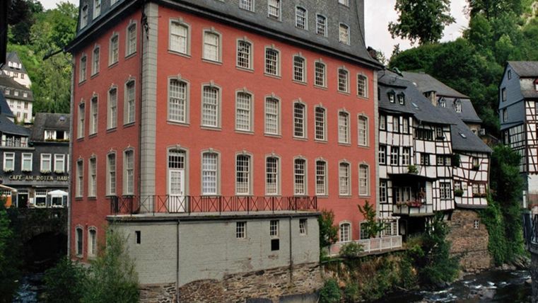 Ein historisches Gebäude mit roter Fassade neben einem kleinen Fluss. Im Hintergrund sind weitere malerische Häuser zu sehen, umgeben von grünen Bäumen.