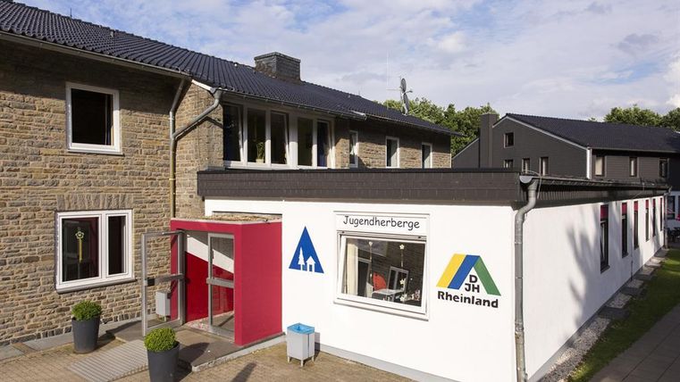 Ein modernes Jugendhaus mit zwei Gebäuden und einem freundlichen Eingangsbereich. Die Fassade ist teilweise weiß und zeigt das Logo der Jugendherbergen Rheinland.
