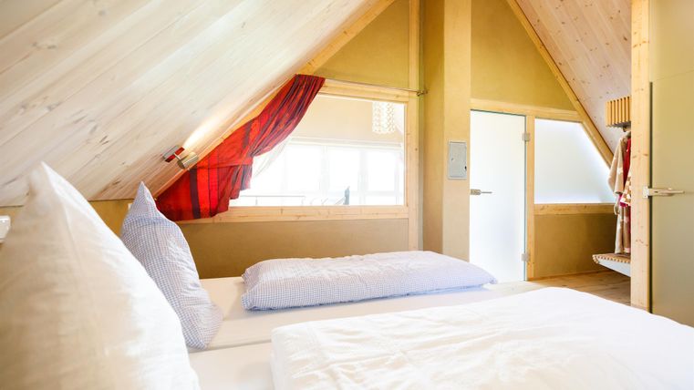 Ein gemütliches Schlafzimmer mit schrägen Wänden und Holzverkleidung. Die Atmosphäre ist hell und einladend, mit großen Fenstern und modernen Möbeln.