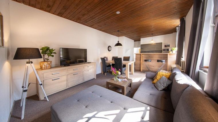 Ein modernes Wohnzimmer mit einer gemütlichen Couch und einem Fernseher. Der Raum hat Holzdecken und ist hell mit einer offenen Küchenzeile.