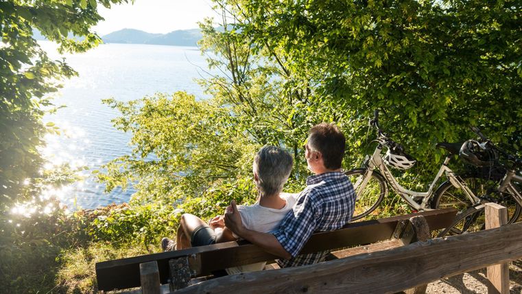 Ein Paar sitzt auf einer Bank und genießt die Aussicht auf das Wasser und die Natur. Zwei Fahrräder stehen neben der Bank.