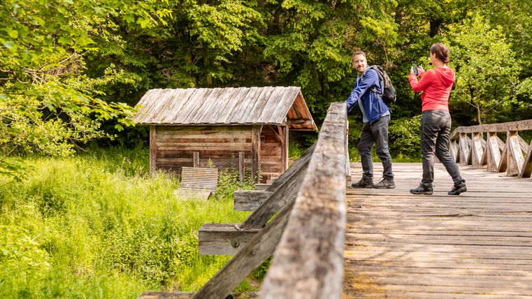Zwei Personen auf einer Holzbrücke vor einer Holzhütte im Grünen.