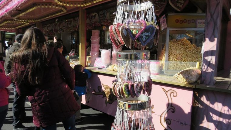 Ein bunter Verkaufsstand auf einer Festveranstaltung mit großen Lebkuchenherzen und Zuckerwatte. Die Menschen stehen in einer Reihe und schauen sich die Süßigkeiten an.