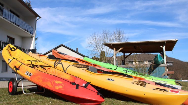 Mehrere farbige Kajaks stehen auf einer Wiese in der Nähe eines Hauses. Im Hintergrund sieht man einen klaren Himmel und eine Holzüberdachung.