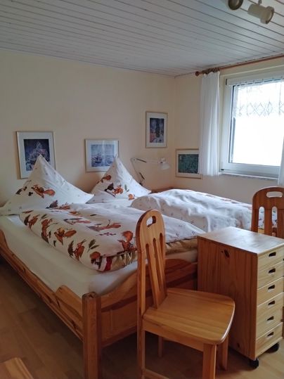 Ein einfaches Schlafzimmer mit zwei Betten und Holzstühlen. Die Wände sind mit Bildern dekoriert und ein Fenster lässt viel Licht herein.