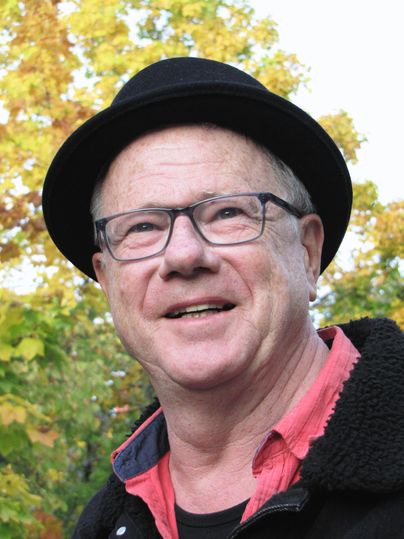 Ein lächelnder Mann mit Brille und schwarzem Hut steht vor einer grünen Kulisse. Er trägt ein rotes Hemd und wirkt freundlich.