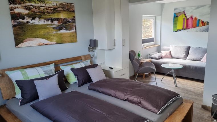 Ein modernes Schlafzimmer mit zwei Betten und bunten Kissen. Im Hintergrund gibt es einen Wohnbereich mit einem Sofa und einem Tisch.