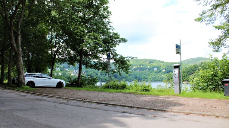 Eine ruhige Straße mit Bäumen und einem weißen Auto. Im Hintergrund ist ein See und hügelige Landschaft zu sehen.