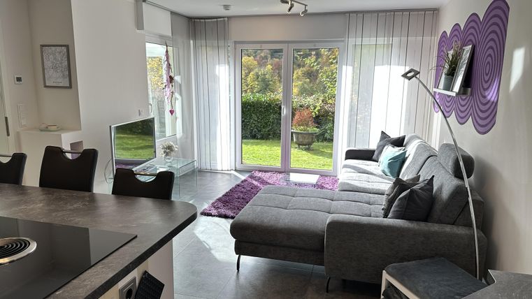 Ein modernes Wohnzimmer mit einem grauen Sofa und großen Fenstern, die den Blick auf den Garten freigeben. Der Raum ist hell und freundlich gestaltet, mit einem stylischen Teppich und dekorativen Elementen.