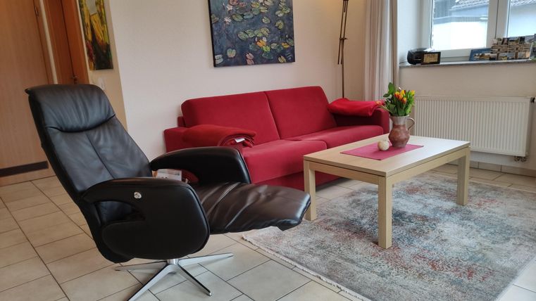 Ein gemütliches Wohnzimmer mit einem roten Sofa und einem schwarzen Drehstuhl. Auf dem Tisch steht eine Vase mit Blumen und im Hintergrund hängt ein Gemälde.