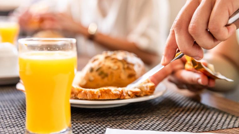 Ein Glas Orangensaft steht auf dem Tisch neben einem gebutterten Brötchen. Eine Hand schneidet das Brötchen, während im Hintergrund weitere Personen zu sehen sind.