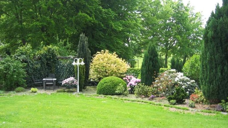 Ein gepflegter Garten mit bunten Blumen und grünen Sträuchern. Im Hintergrund steht eine Bank unter schattenspendenden Bäumen.