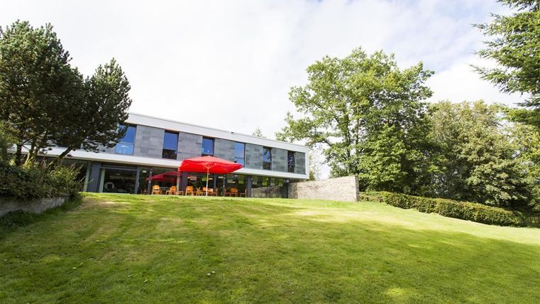 Ein modernes Haus auf einer grünen Wiese, umgeben von Bäumen. Im Vordergrund steht ein roter Sonnenschirm über einem Sitzbereich.