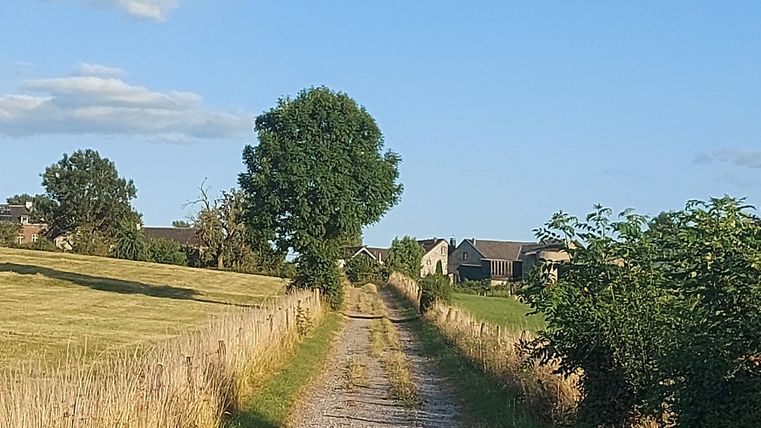Ein Schotterweg, der durch eine ländliche Landschaft führt. Am Ende des Weges sind einige Häuser sichtbar, unter einem klaren blauen Himmel.