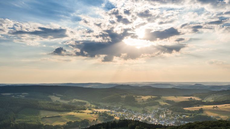 Landschaftsansicht von Neroth mit bewaldeten Hügeln und einem Dorf im Tal, unter einem Himmel mit Wolken und Sonnenstrahlen.