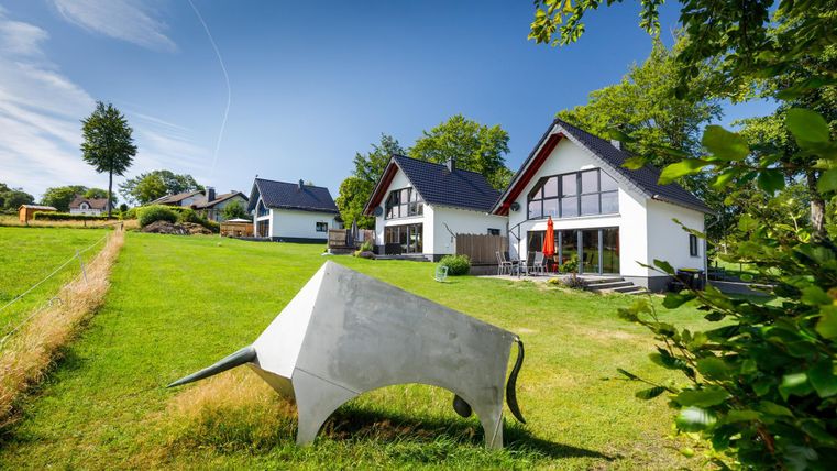 Ein modernes Haus mit großen Fenstern steht inmitten einer grünen Wiese. Im Vordergrund befindet sich eine auffällige, skulpturale Kuh aus Beton.