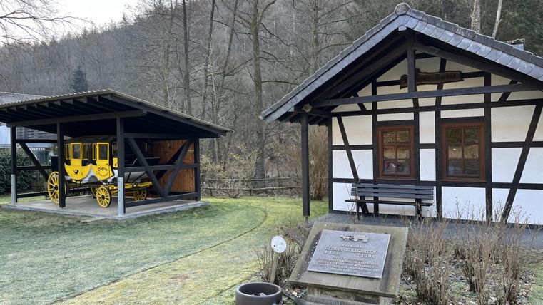 Ein historisches Gebäude mit Holzrahmen und großen Fenstern steht neben einer überdachten Fläche, in der eine gelbe Kutsche ausgestellt ist. Die Umgebung ist von Bäumen umgeben und es ist eine grüne Wiese sichtbar.