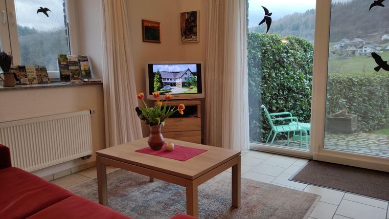 Ein gemütliches Wohnzimmer mit einem roten Sofa und einem Holztisch. Durch die Fenster sieht man eine grüne Terrasse und die umliegende Landschaft.