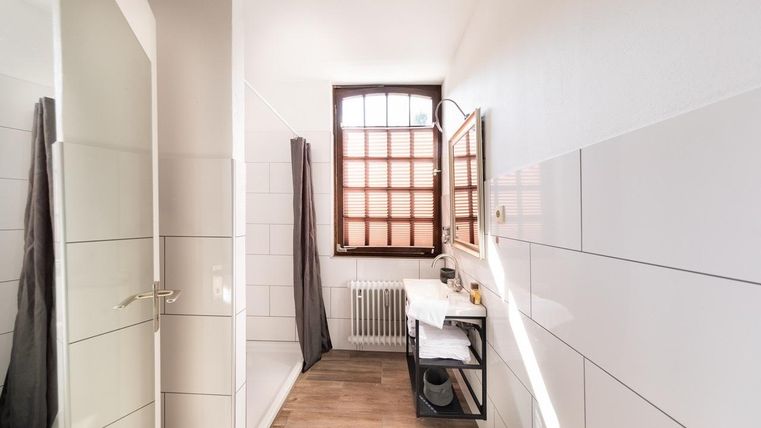 Ein helles Badezimmer mit weißen Fliesen und Holzfußboden. Es gibt eine Dusche, ein Waschbecken und ein Fenster mit Holzjalousien.