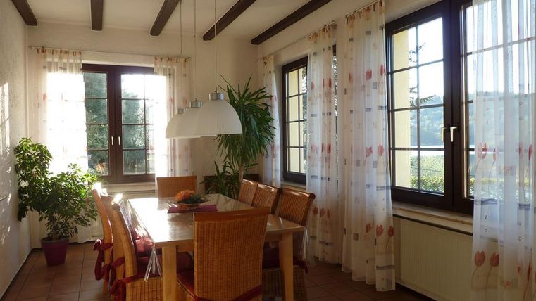 Ein helles Esszimmer mit einem großen Tisch und Stühlen. Die Fenster sind mit Vorhängen dekoriert, und eine Pflanze steht in der Ecke.